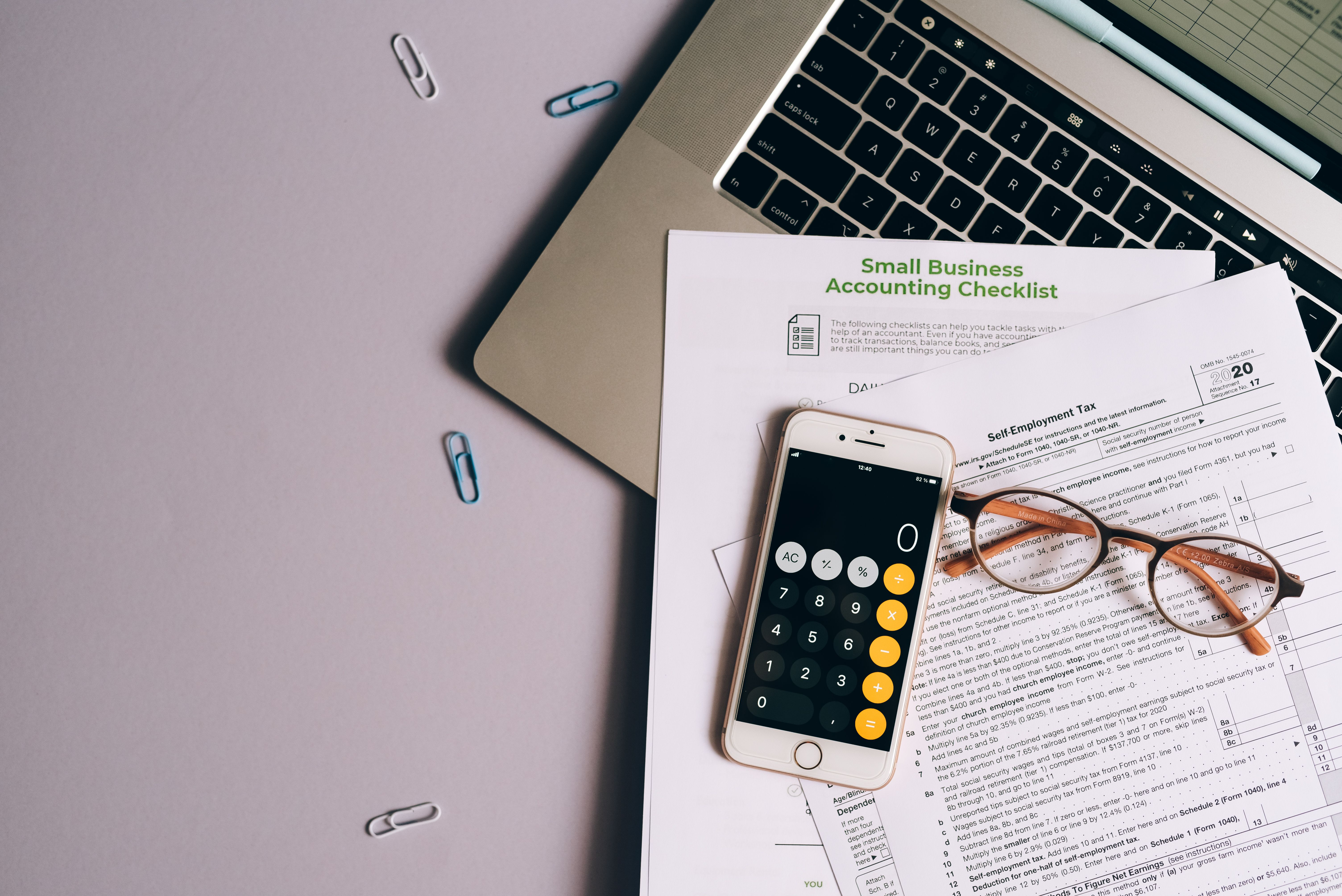 Small-business tax forms, smartphone, and glasses arranged on a desk for tax preparation and recordkeeping.